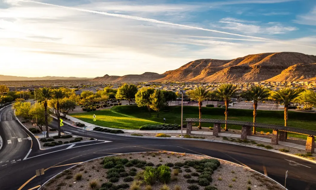 Summerlin_Sustainability-breathe-easier A roundabout surrounded by desert landscaping leads to a lush green park with palm trees; in the background, suburban houses stretch toward sunlit desert hills under a blue sky with wispy clouds.