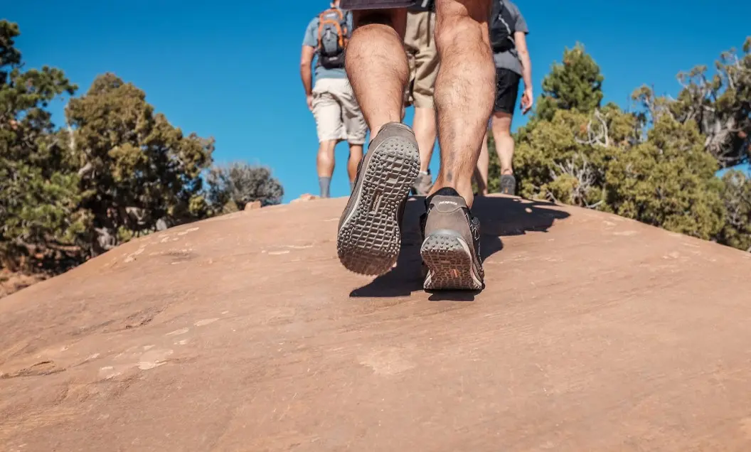 Summerlin_Sustainability-stewardship-action Close-up of hikers’ legs and shoes as they walk up a rocky slope, with blue sky and green trees in the background. The focus is on the soles of the shoes and the textured rock surface.