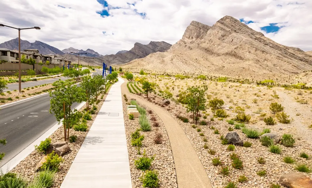 A wide sidewalk and a parallel walking path run through a landscaped desert area with small green plants, near a roadway and houses, with rocky mountains and a partly cloudy sky in the background.