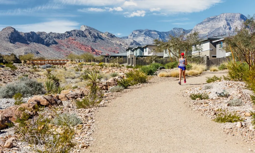 A person in athletic wear jogs on a gravel trail through a desert landscape with shrubs, mountains in the background, and houses lining one side under a partly cloudy sky.