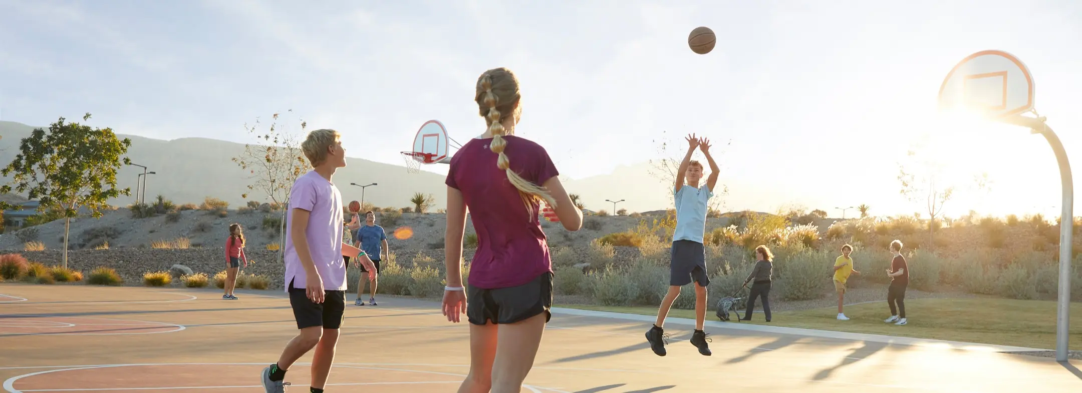 A group of kids play basketball outdoors on a sunny day, with one child jumping to shoot the ball while others watch and stand on the court. There are trees, mountains, and more children in the background.