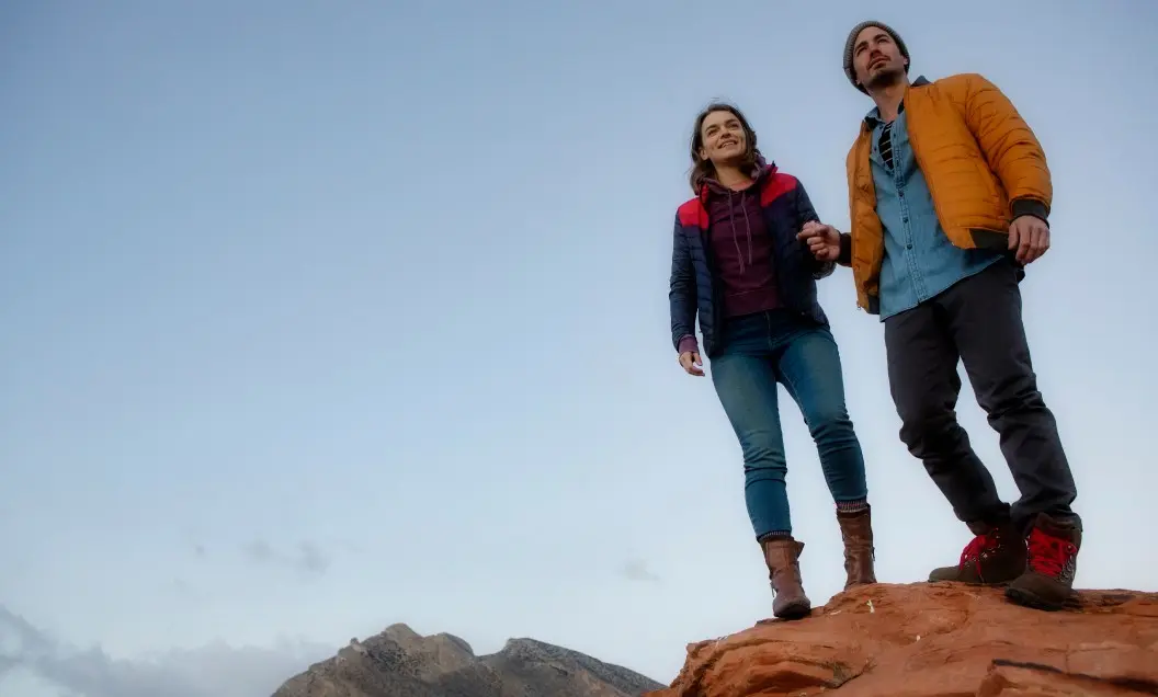 A man and woman wearing jackets and boots stand together on a rocky ledge, holding hands and smiling, with mountains and a clear sky in the background.