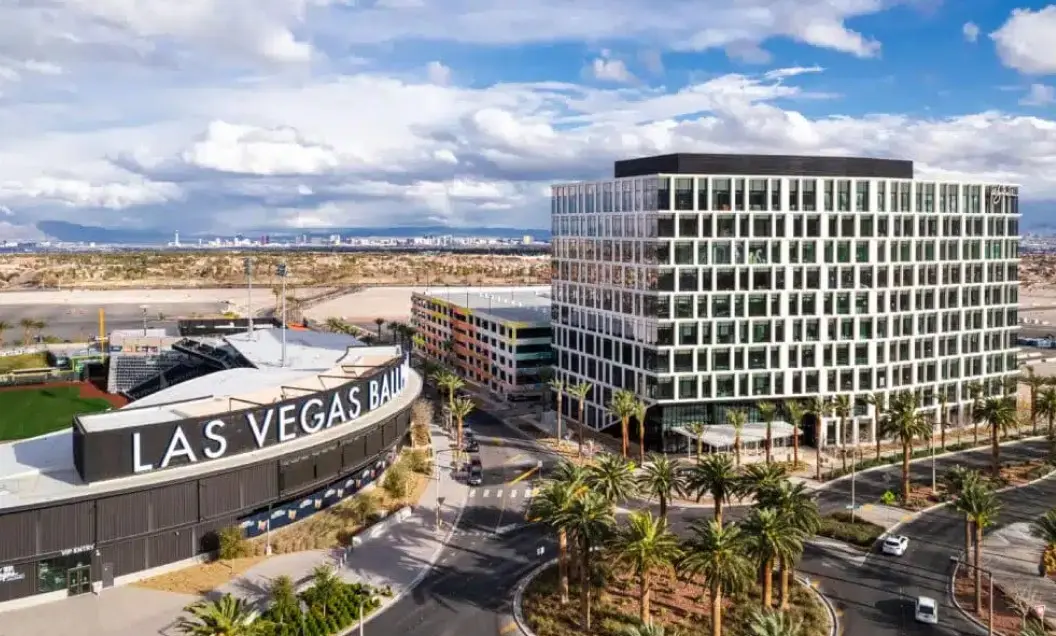 Summerlin_Why-Business Aerial view of Las Vegas Ballpark and a modern office building surrounded by palm trees, with desert landscape and mountains visible in the background under a partly cloudy sky.