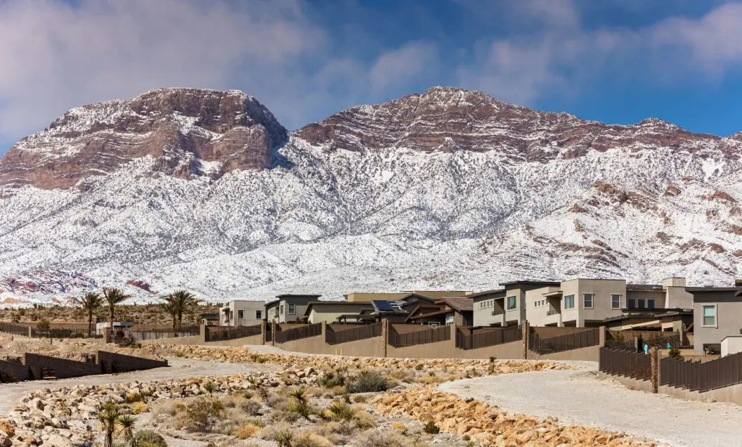 Summerlin_Why-Cool-Factor Modern houses line a suburban neighborhood with desert landscaping in the foreground, set against a backdrop of large, rocky mountains dusted with snow under a partly cloudy blue sky.