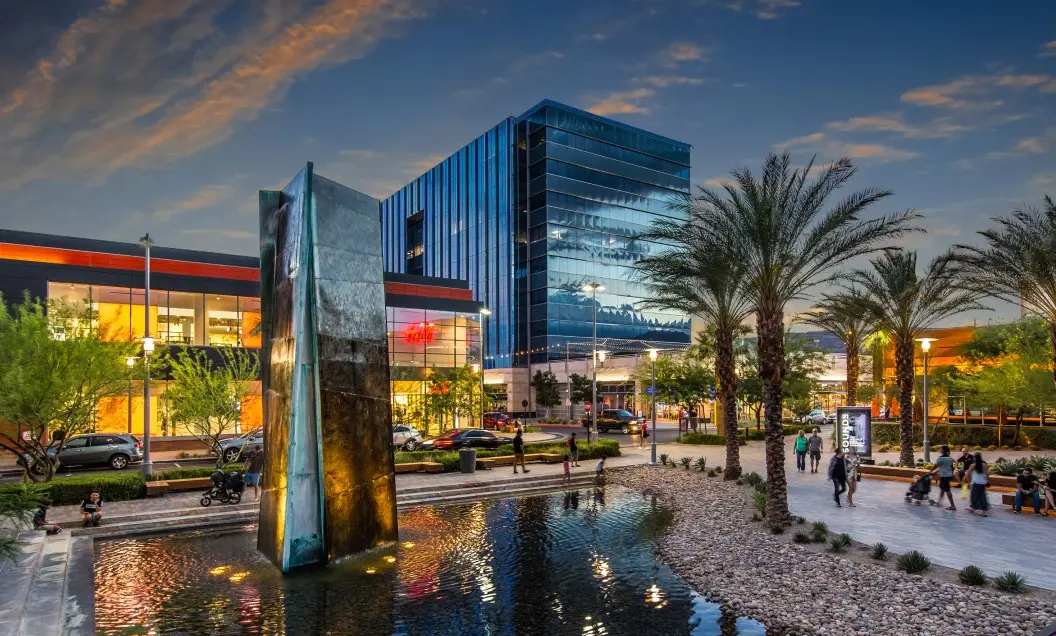Summerlin_Why-Downtown A modern plaza at dusk features a tall, angular water sculpture beside a reflecting pool, surrounded by palm trees, glass office buildings, shops, and people walking along tree-lined paths.
