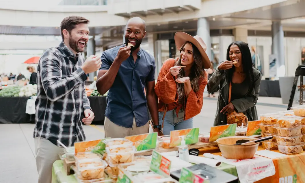 Summerlin_Why-FOMO Four people smile and laugh as they sample food at an outdoor market stall filled with dips and chips, enjoying their time together in a lively, open-air setting.
