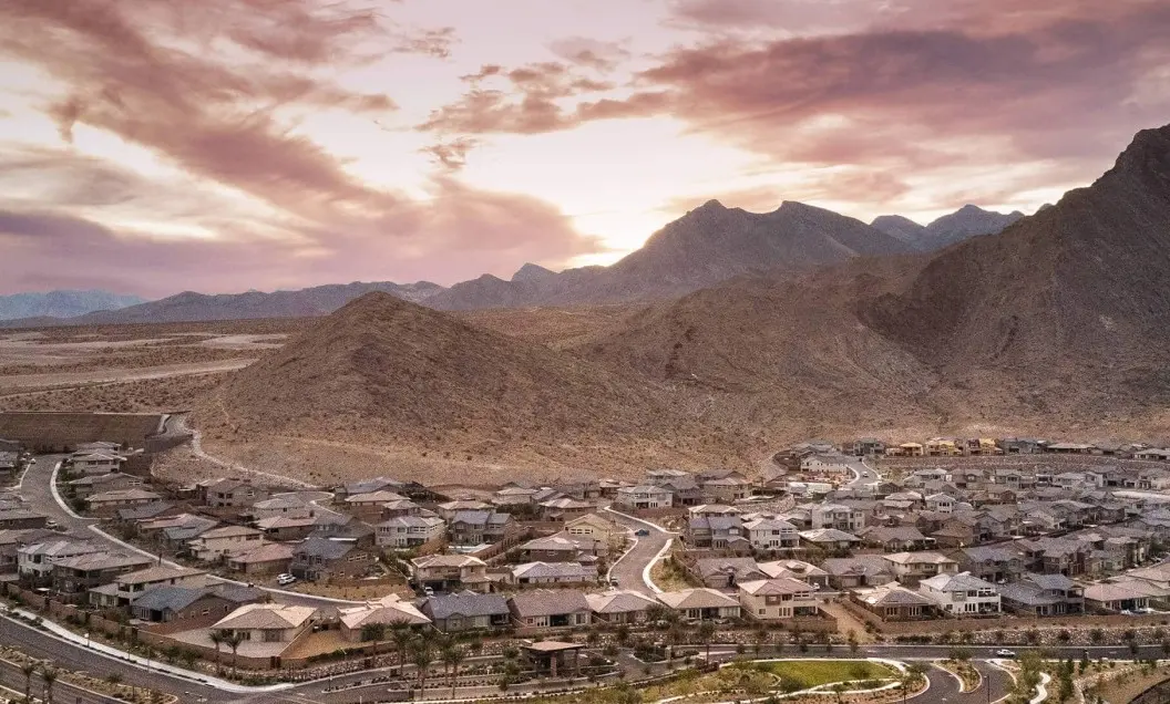Summerlin_Why-Leading-Builders A residential neighborhood with rows of houses sits at the base of rocky mountains under a partly cloudy, colorful sunset sky. Curved streets wind through the community, with desert terrain in the background.
