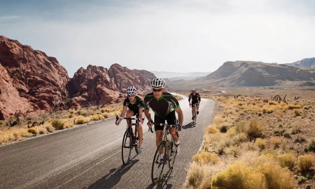 Summerlin_Why-Location Three cyclists ride on a paved road through a desert landscape with rocky hills and sparse vegetation under a bright sky.