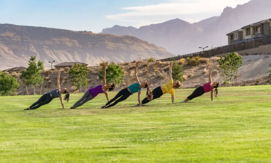 Summerlin_Why-Well-Being Five people exercise outdoors on a grassy field, performing side plank poses in a row. They are dressed in athletic wear, with mountains and houses visible in the background under a blue sky.
