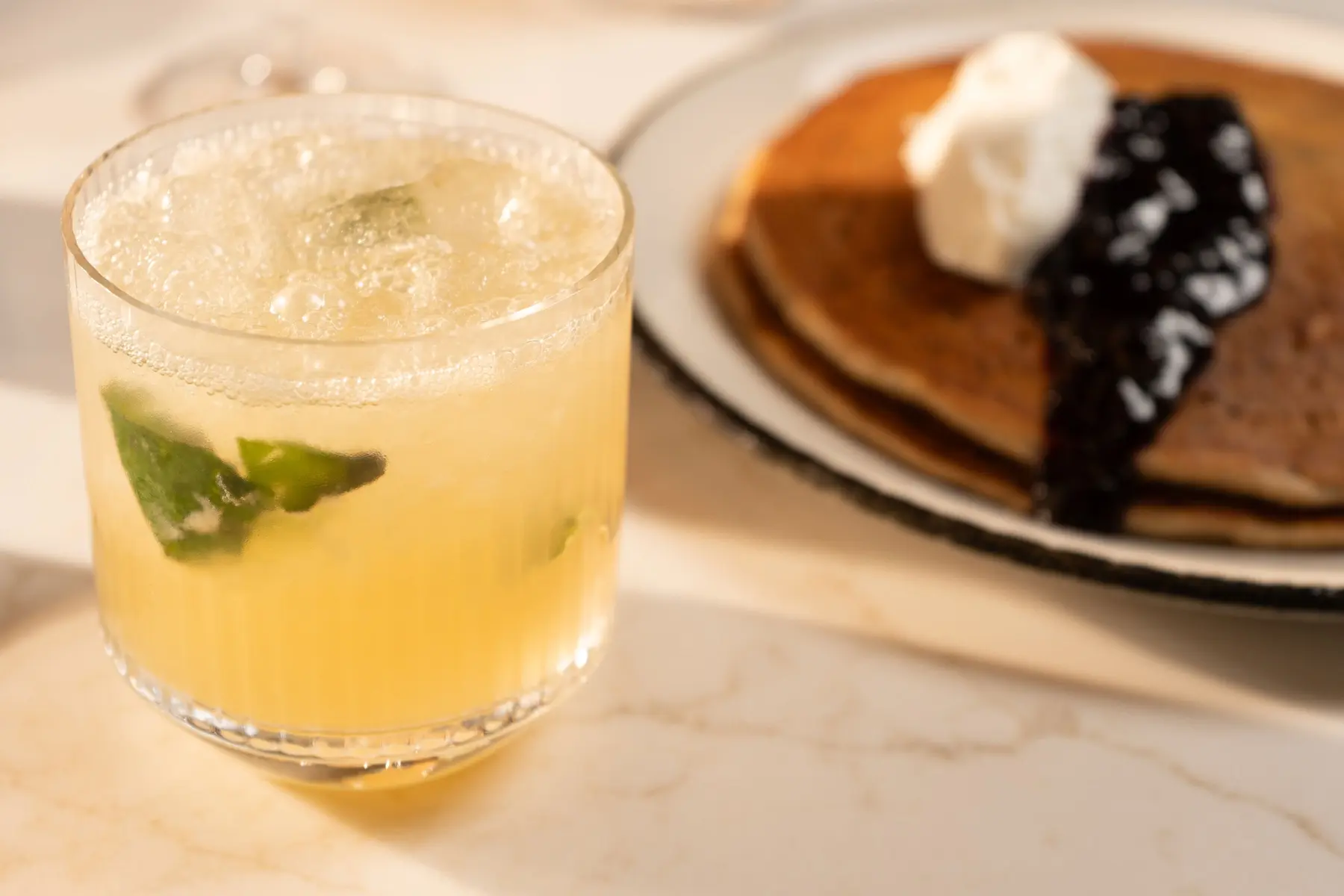 A glass of a light yellow cocktail with ice and mint sits on a table next to a plate of pancakes topped with whipped cream and dark berry sauce.