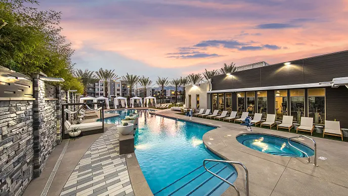 Tanger Apartments-1 2_cropped Luxury outdoor pool area at sunset with lounge chairs, cabanas, a hot tub, palm trees, and modern apartment buildings in the background. The sky has pink and purple hues.