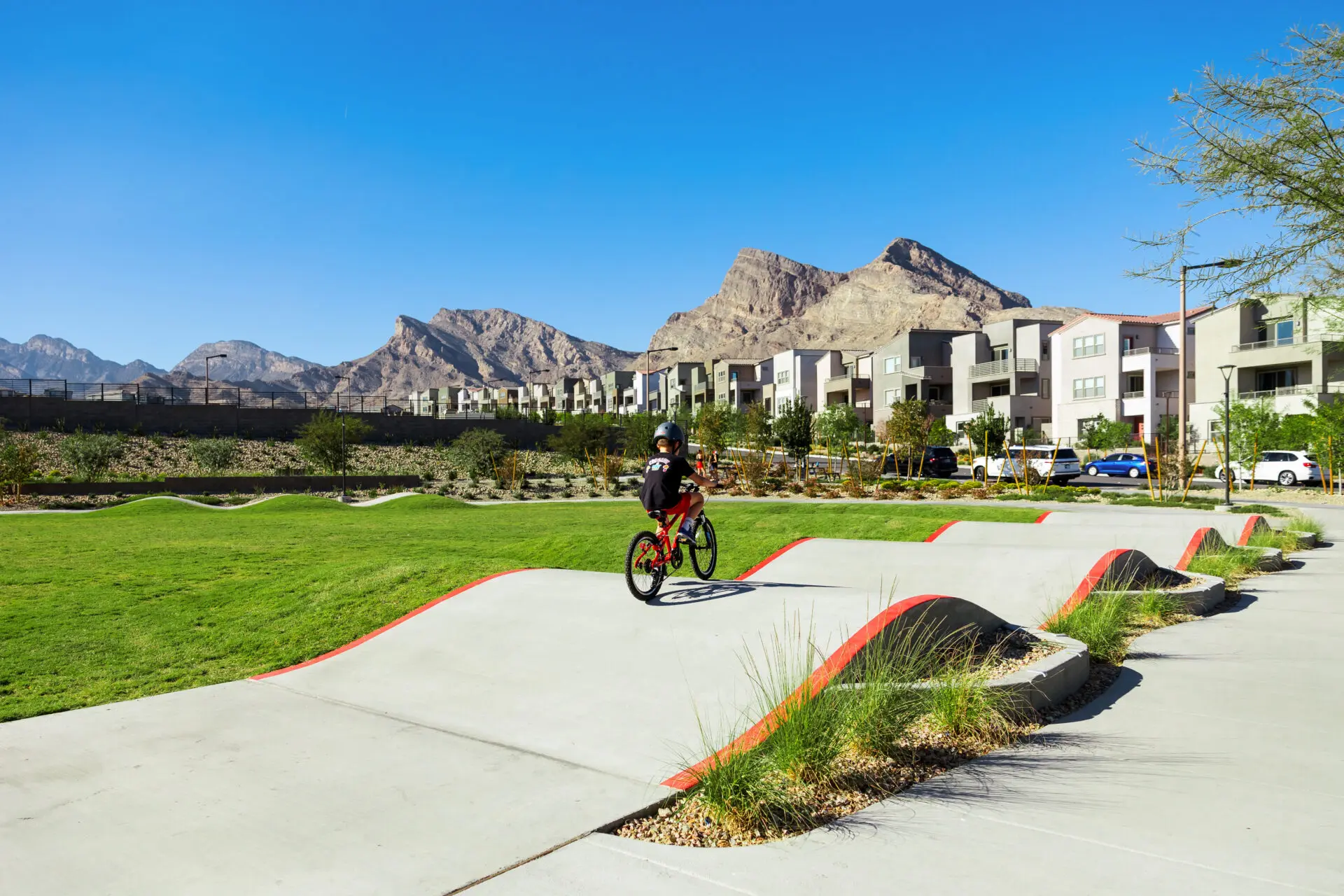 A person wearing a helmet rides a bike on a winding concrete path with small hills, surrounded by green grass, modern apartments, and mountains under a clear blue sky.