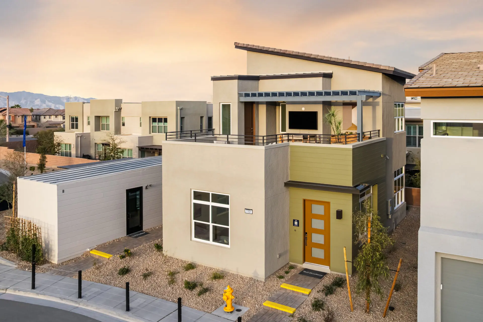The Loughton Modern two-story house with geometric design, large windows, a rooftop balcony, and a yellow front door. The property has desert landscaping and is located on a corner lot in a suburban neighborhood at sunset.