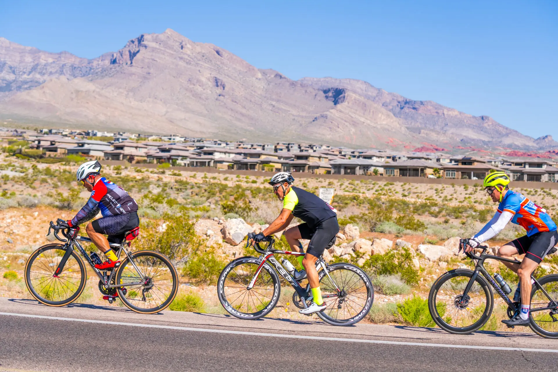 Three cyclists in colorful gear ride on a road through a desert landscape with mountains and houses in the background under a clear blue sky.