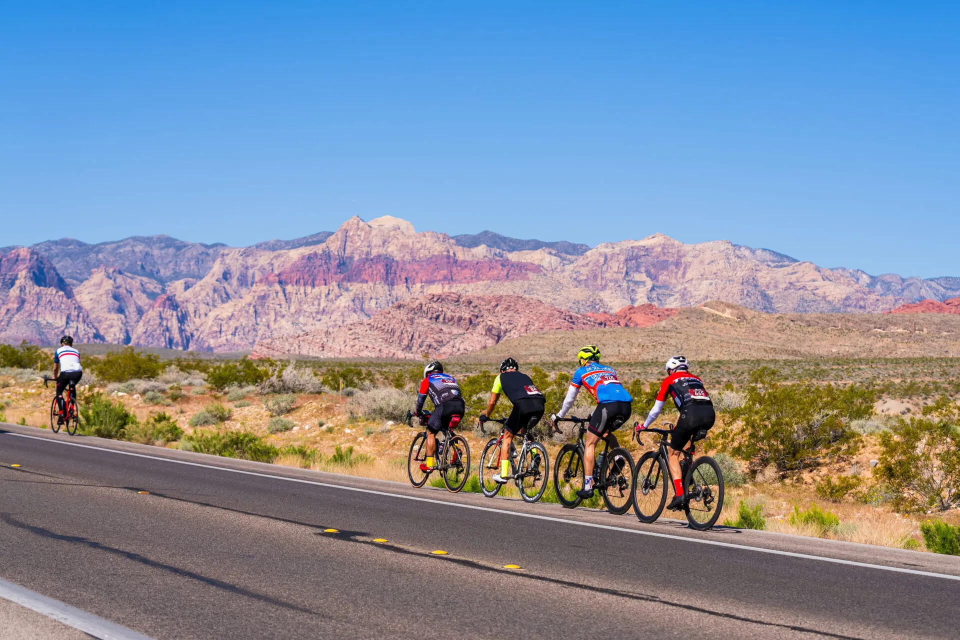 A group of cyclists ride along a desert highway with colorful mountains in the distance under a clear blue sky.