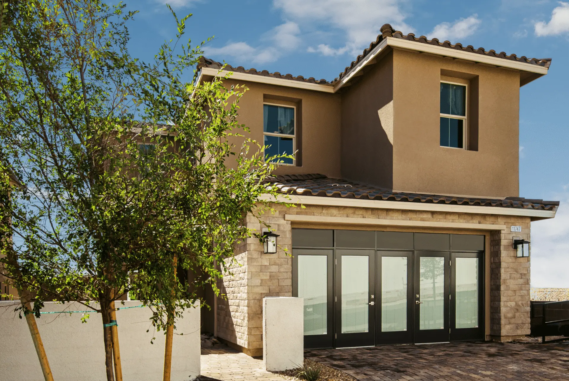Two-story modern house with tan stucco, stone accents, a dark tiled roof, and large glass doors. A small tree and paved walkway are in the foreground, under a bright blue sky with scattered clouds.