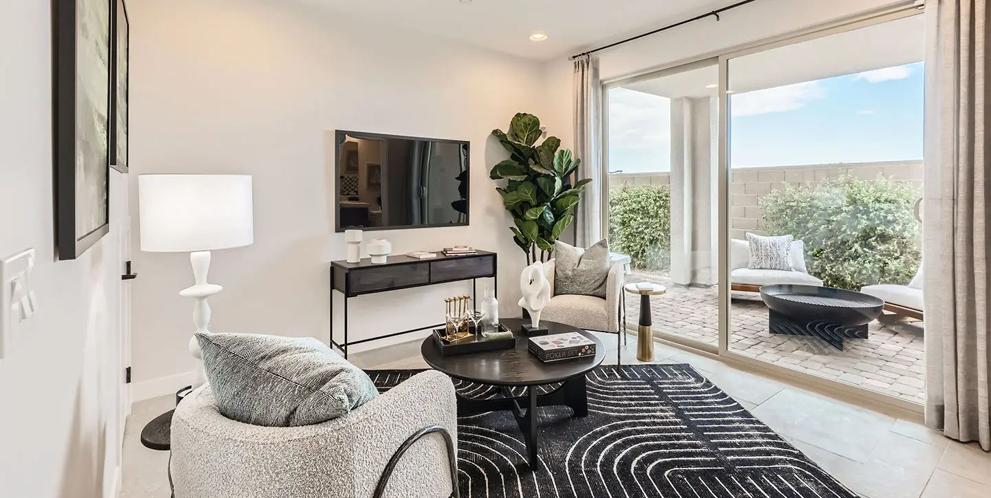 Modern living room with two gray chairs, a black round coffee table on a black-and-white rug, a large TV on a console, a tall plant, and sliding glass doors leading to a patio with outdoor seating and a brick wall.