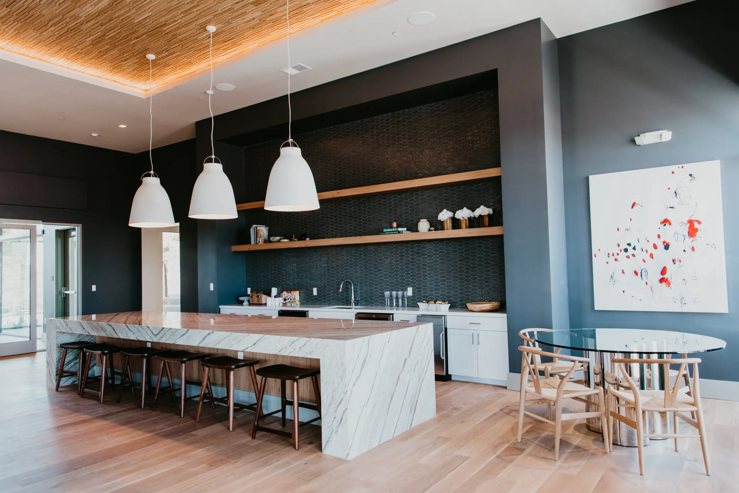 b7a9737f67c4e63c7c83ab8f18bc10fa353f432f A modern kitchen with a large marble island, wooden stools, dark textured walls, open wooden shelves, three white pendant lights, and a round glass dining table with wooden chairs. Abstract art hangs on the wall.