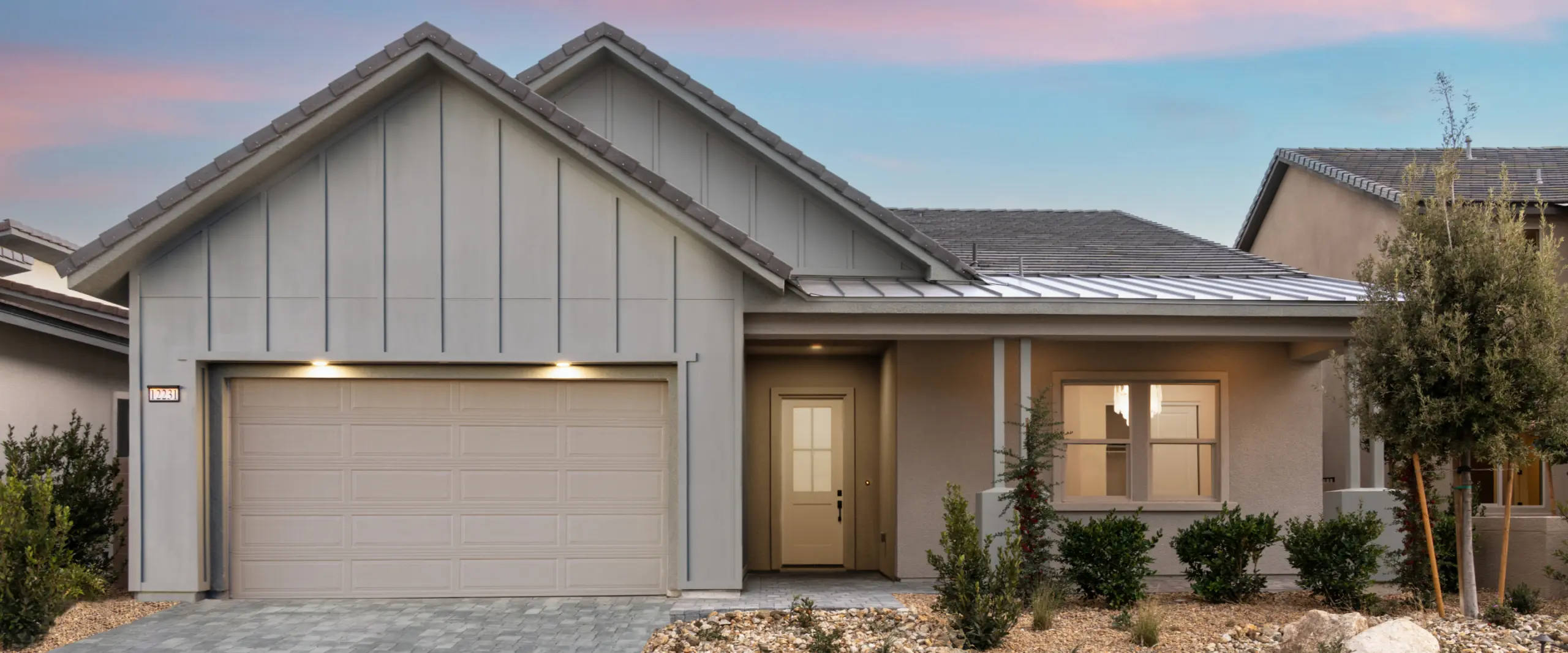 Modern single-story house with a light gray exterior, a double garage, a covered porch, a small front yard with shrubs, and a paved driveway, set under a pink and blue evening sky.