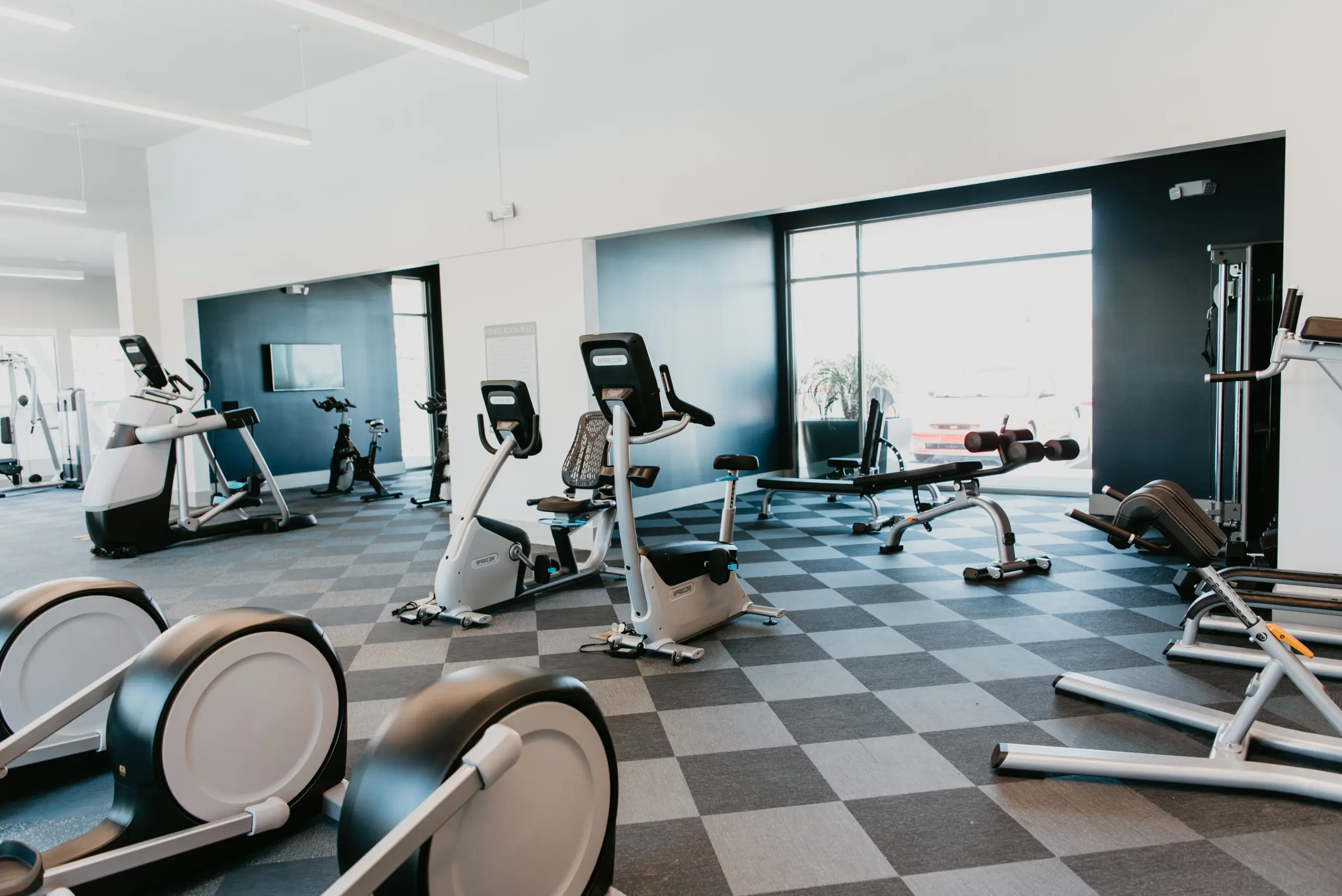 d29209f81a27a90de30b017ab296509d6fe96801 A modern gym with exercise bikes, benches, and various fitness machines on a checkered floor, with large windows letting in natural light.