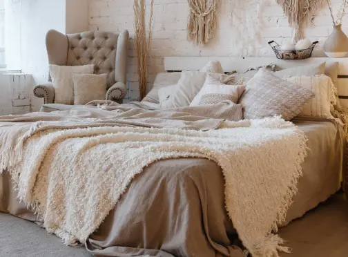 A cozy bedroom with a large bed covered in beige and cream blankets and pillows, a tufted armchair with cushions, and a white brick wall decorated with macrame art and a basket. Warm, neutral tones throughout.