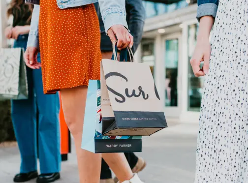 People walking outdoors, carrying shopping bags. One person holds a paper bag that says “Sur,” while another holds multiple bags. The scene suggests a casual shopping outing on a sunny day.