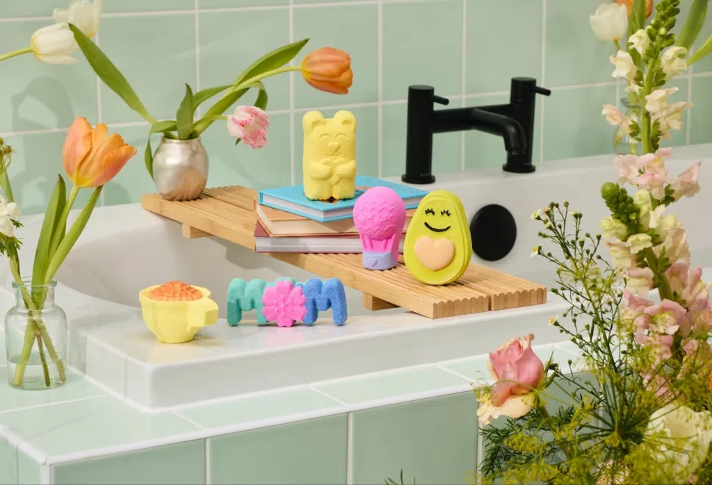A pastel-colored bathroom with a white tub, mint-green tiles, flowers, and playful bath bombs shaped like a bear, ice cream, avocado, and letters spelling MOM arranged on a wooden bath tray.