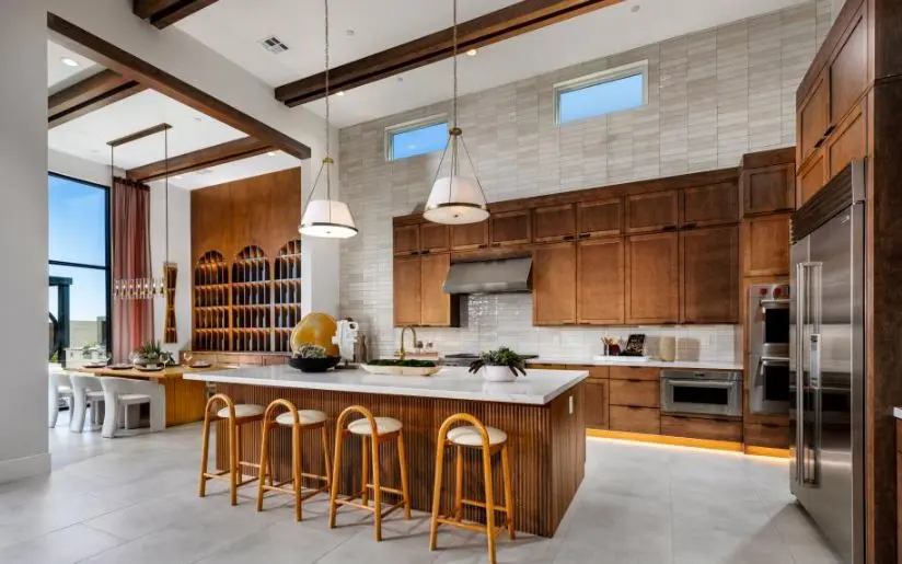 Modern kitchen with wood cabinets, large marble island with four wooden stools, stainless steel appliances, pendant lights, and high ceilings with exposed beams. Dining area is visible to the left with a long table.
