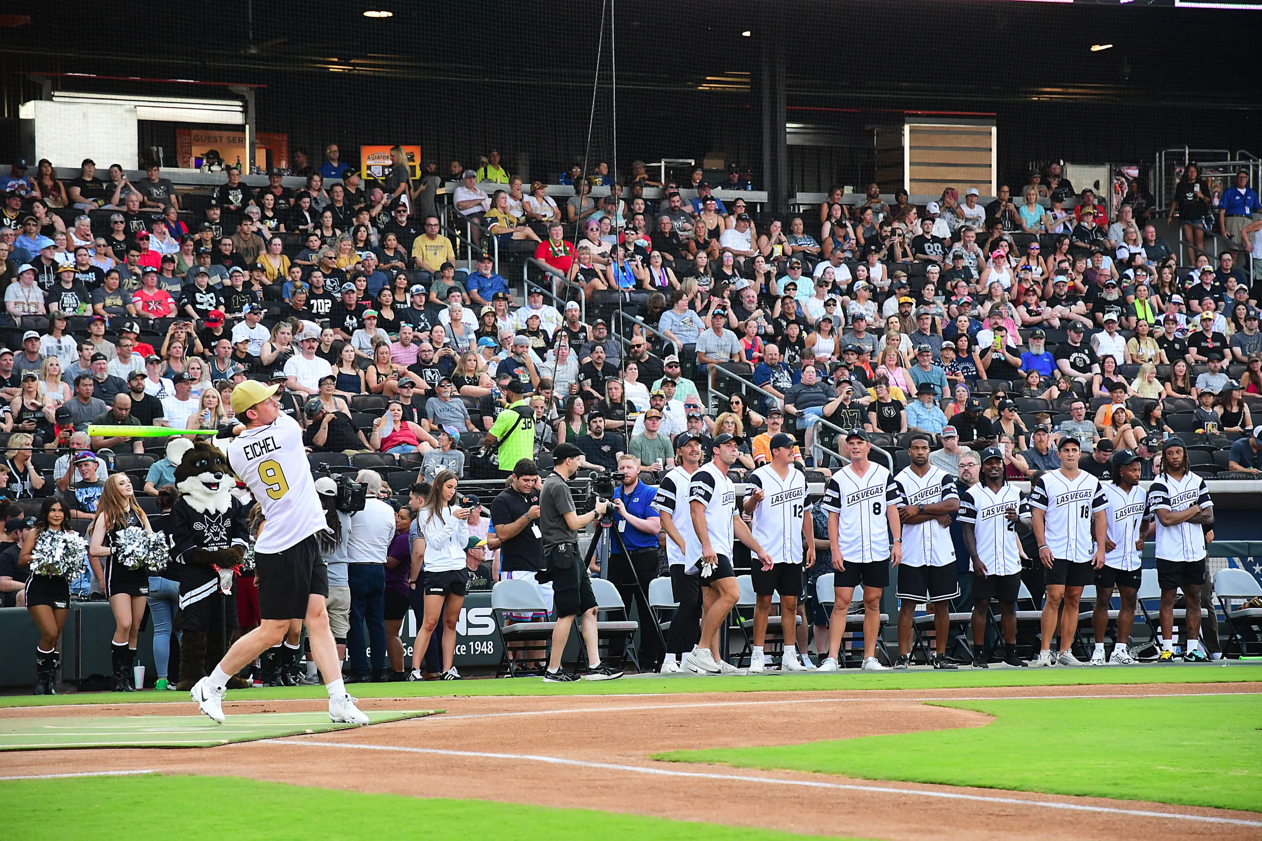A baseball player swings a bat on a field as cheerleaders, teammates in matching jerseys, and a large crowd of spectators watch from the stands. Photographers capture the moment near home plate.