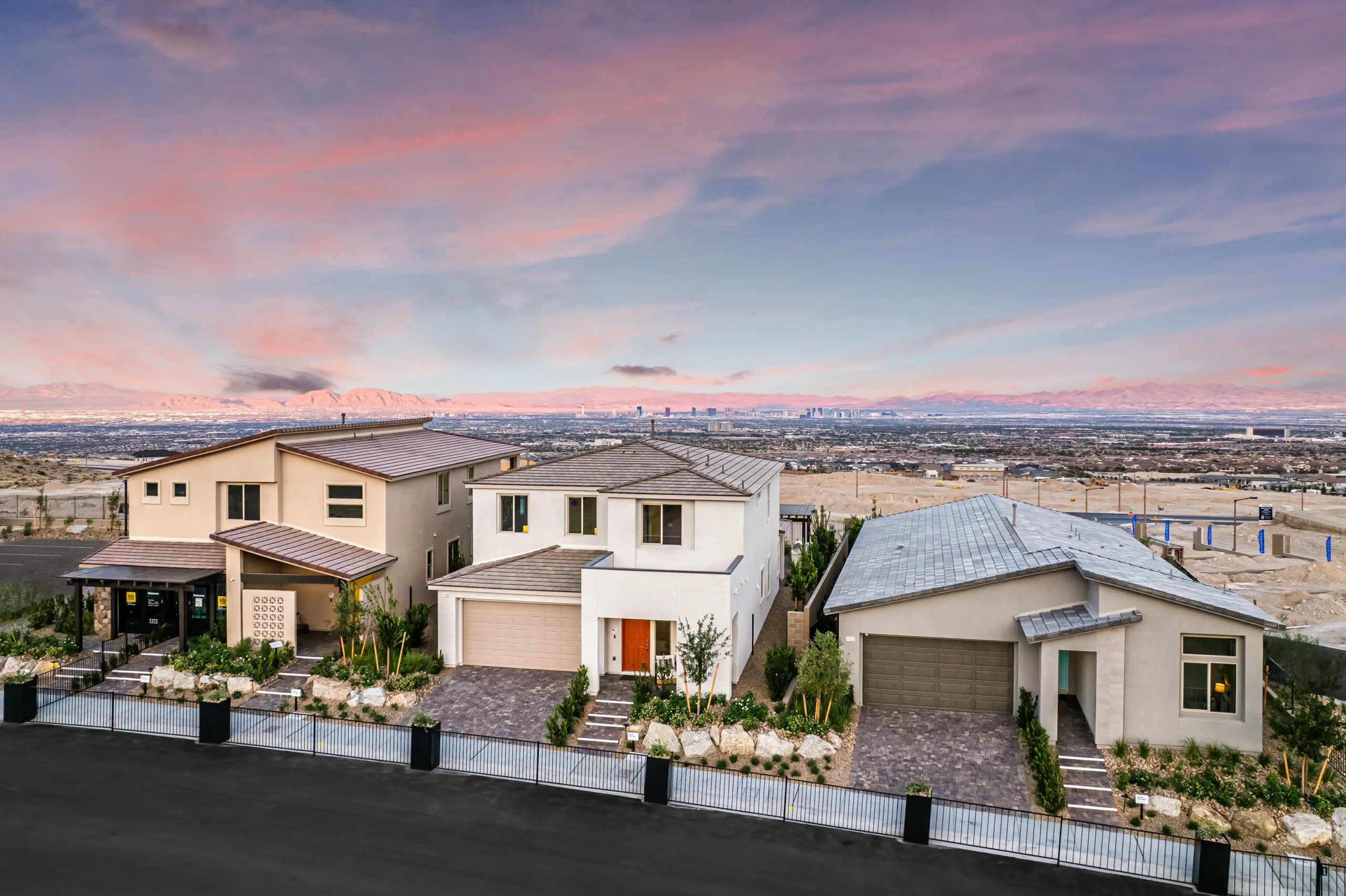 Three modern suburban houses with landscaped yards are lined up on a street, with a scenic city skyline and mountains in the distance under a pink and blue sunset sky.