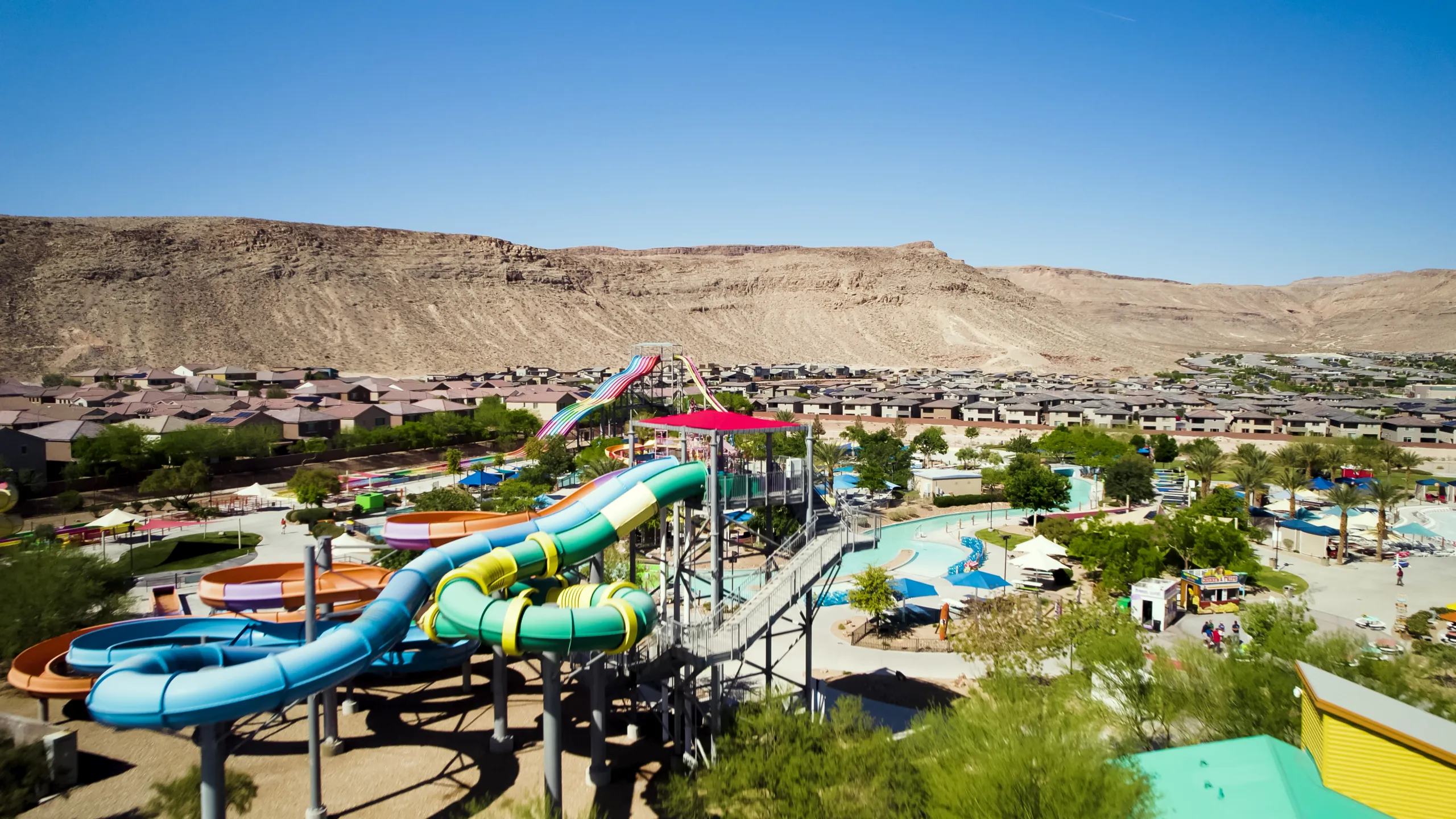 Aerial view of a water park with winding water slides and pools, surrounded by greenery and buildings, set against a backdrop of dry, rocky hills and a clear blue sky.