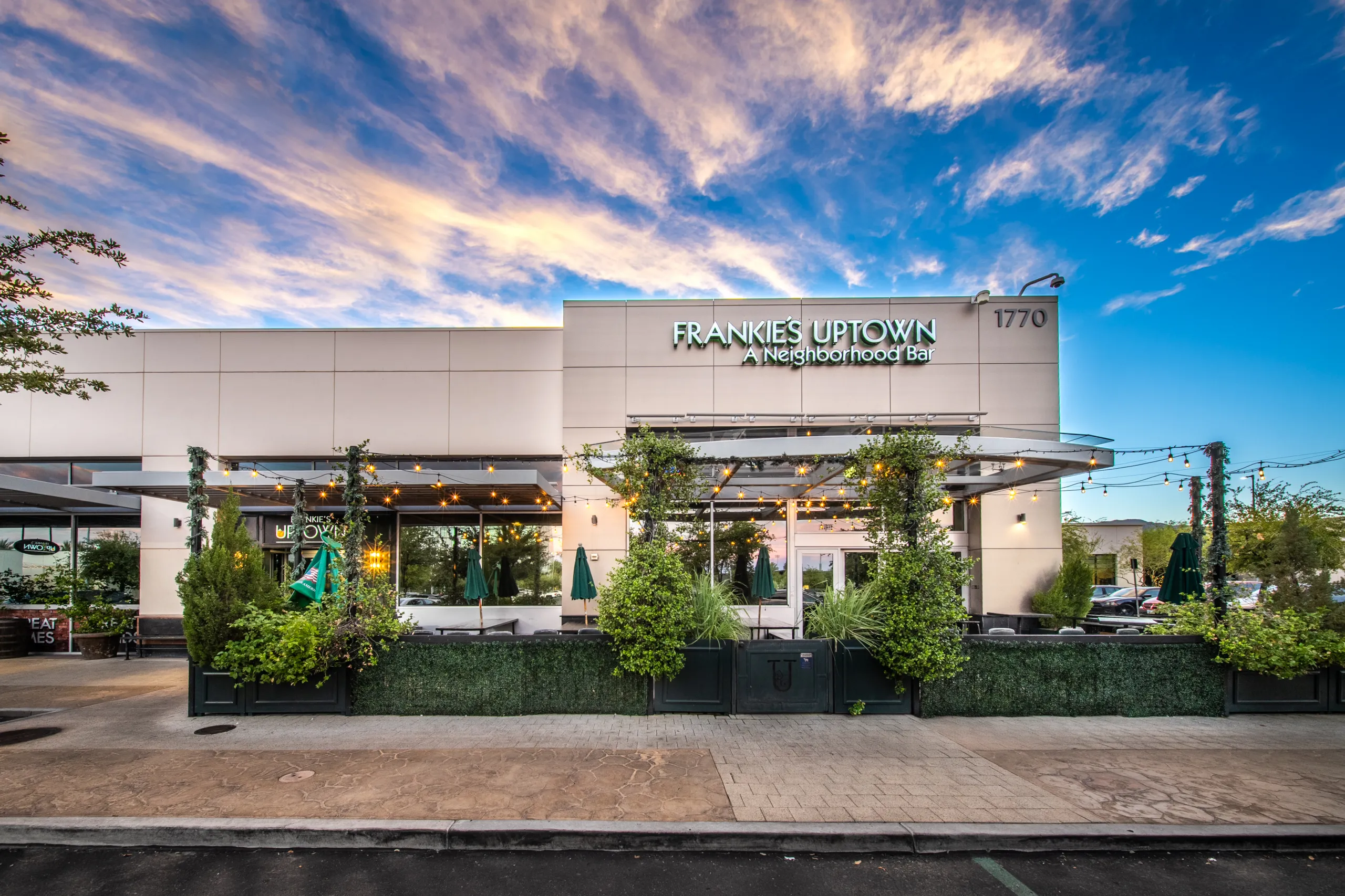 Frankie’s Uptown 2026 (1) Modern bar and restaurant exterior with large windows, greenery, outdoor seating, and a sign reading FRANKIES UPTOWN - A Neighborhood Bar. The sky is blue with scattered clouds at sunset.