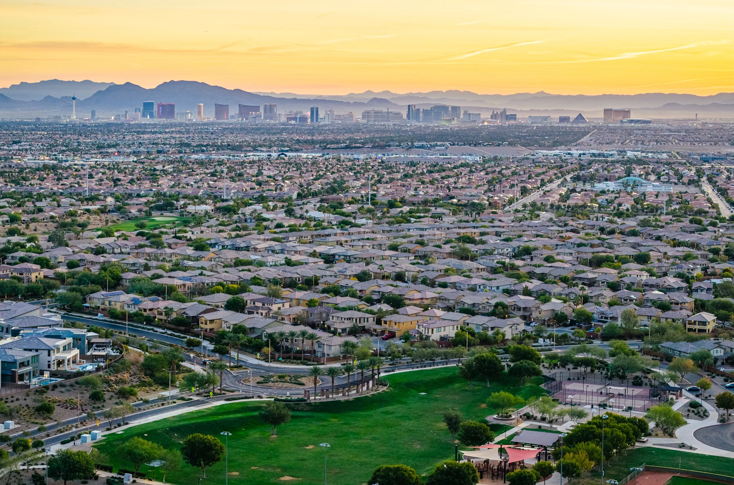 Aerial view of a sprawling suburban neighborhood with green parks in the foreground and the Las Vegas Strip skyline and mountains in the background at sunset.