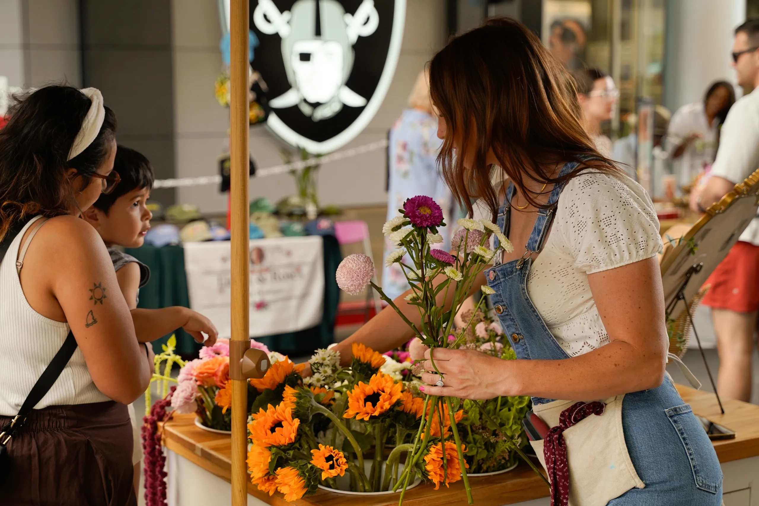 Night Market 2026 A woman in overalls arranges colorful flowers at a market stall, while a child and another woman look on. The table is filled with various vibrant blooms, and people are visible in the background.