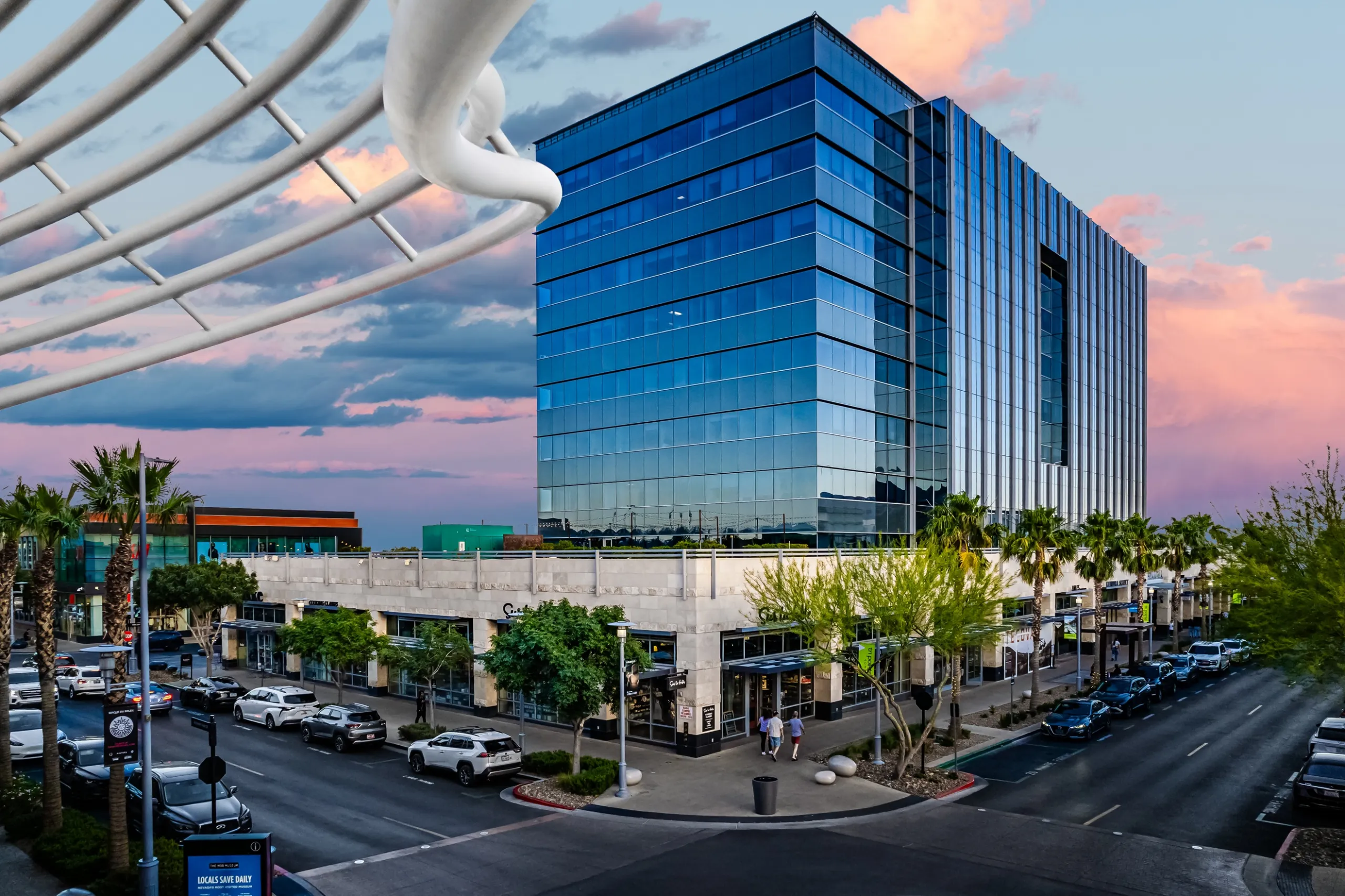 A modern glass office building reflects the sky at sunset, surrounded by palm trees, shops, and parked cars on a busy intersection with people walking along the sidewalks.