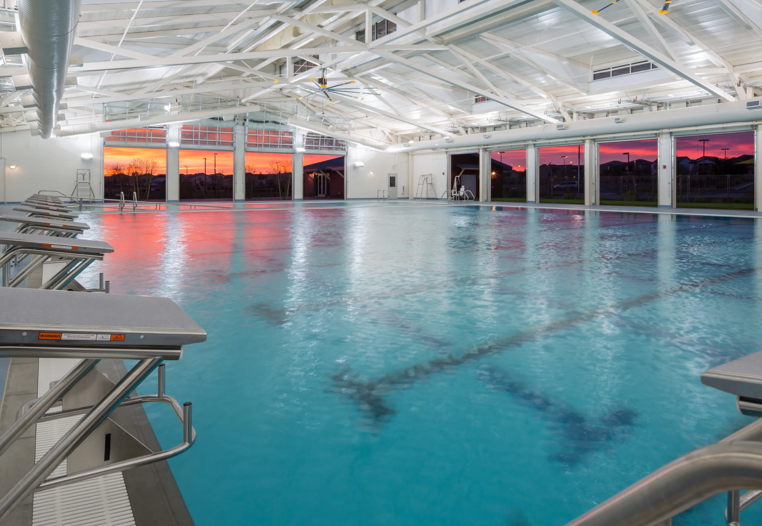 An indoor swimming pool with diving platforms, large windows, and high white ceilings. The sunset outside casts an orange and pink glow across the water and the pool area.