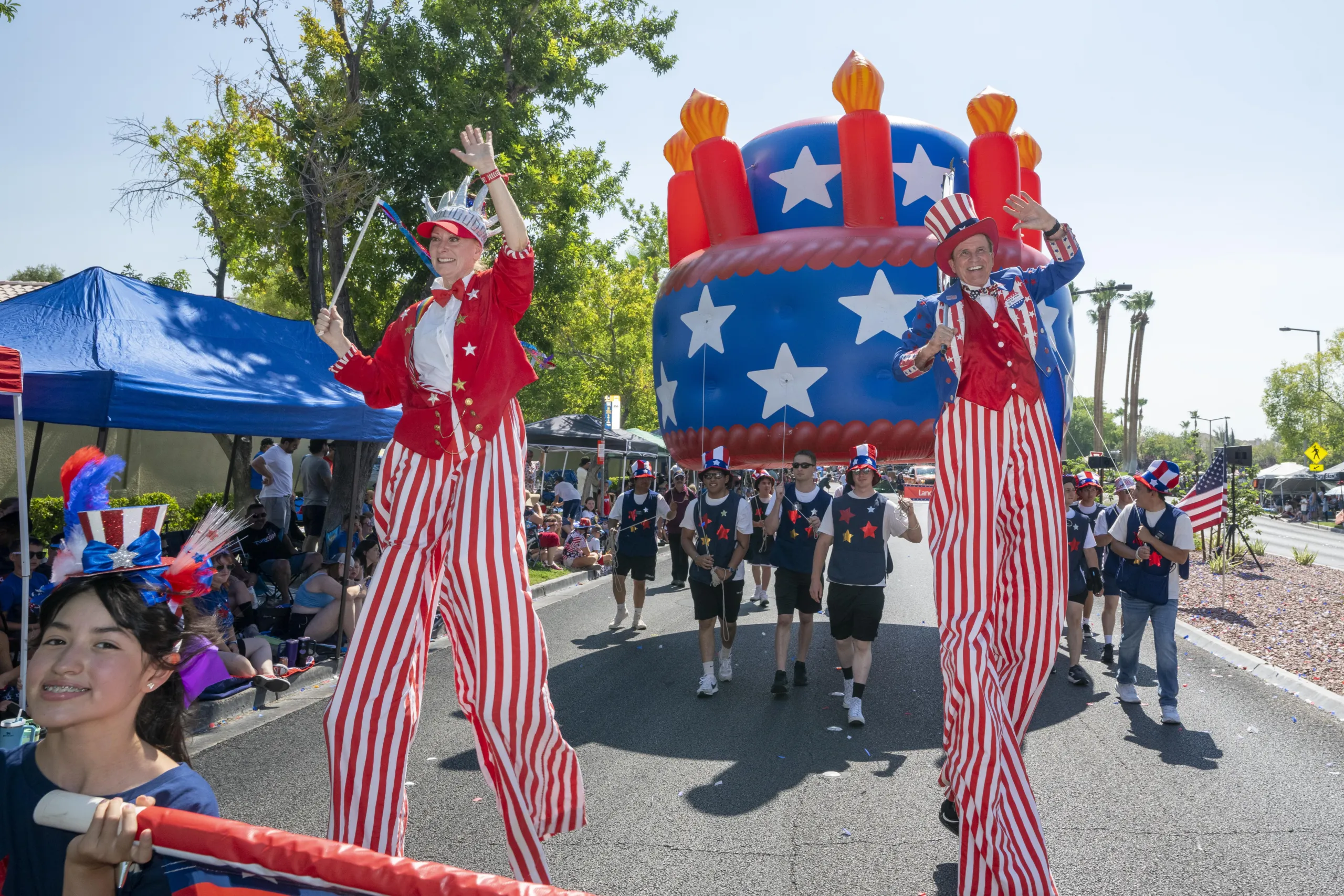 People in patriotic costumes and stilts walk in a parade with a large inflatable cake float decorated in stars and stripes, while spectators watch from the roadside under tents.