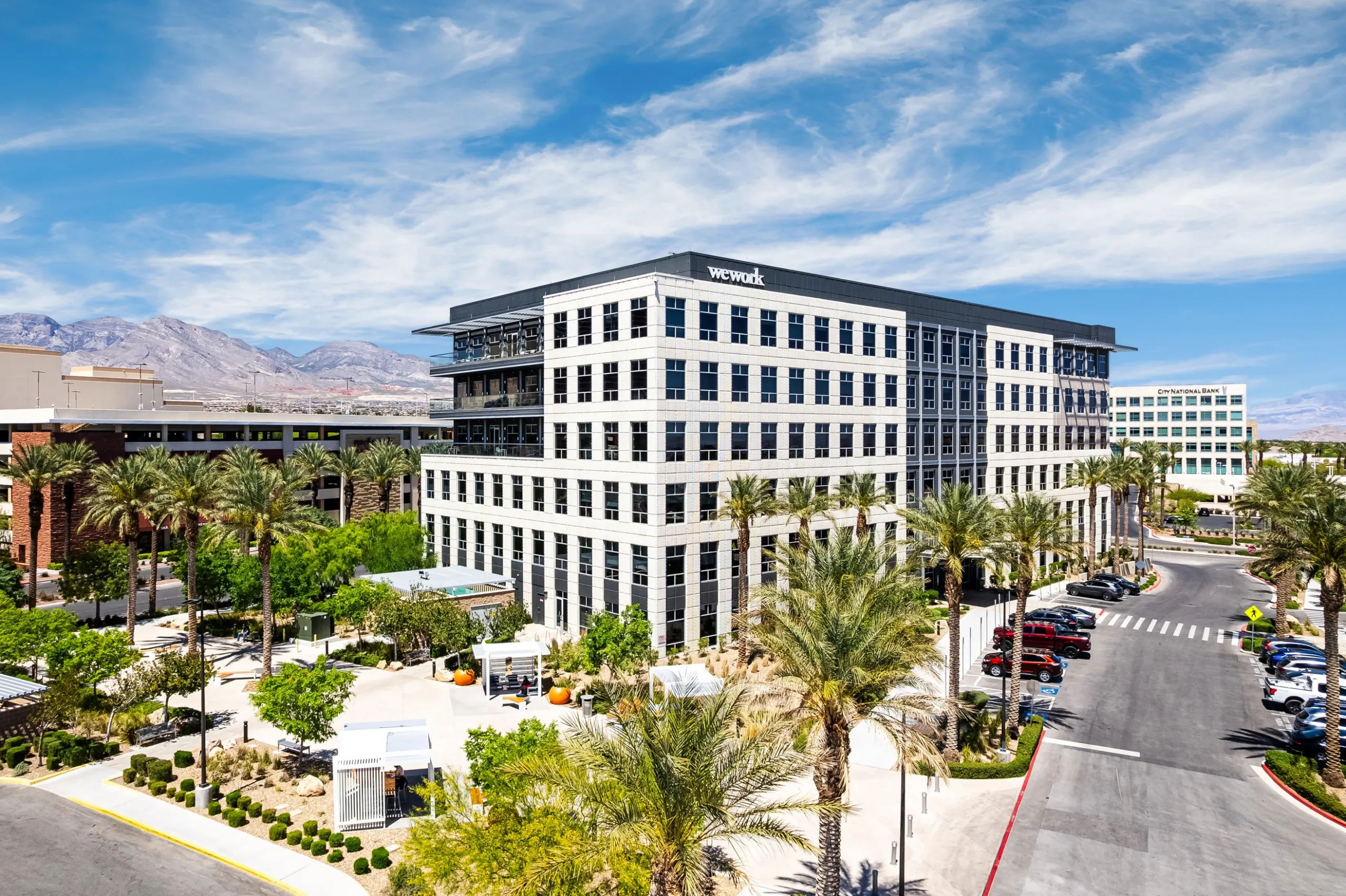 A modern office building with WeWork signage is surrounded by palm trees, parked cars, and landscaping under a bright blue sky with scattered clouds. Mountains are visible in the background.
