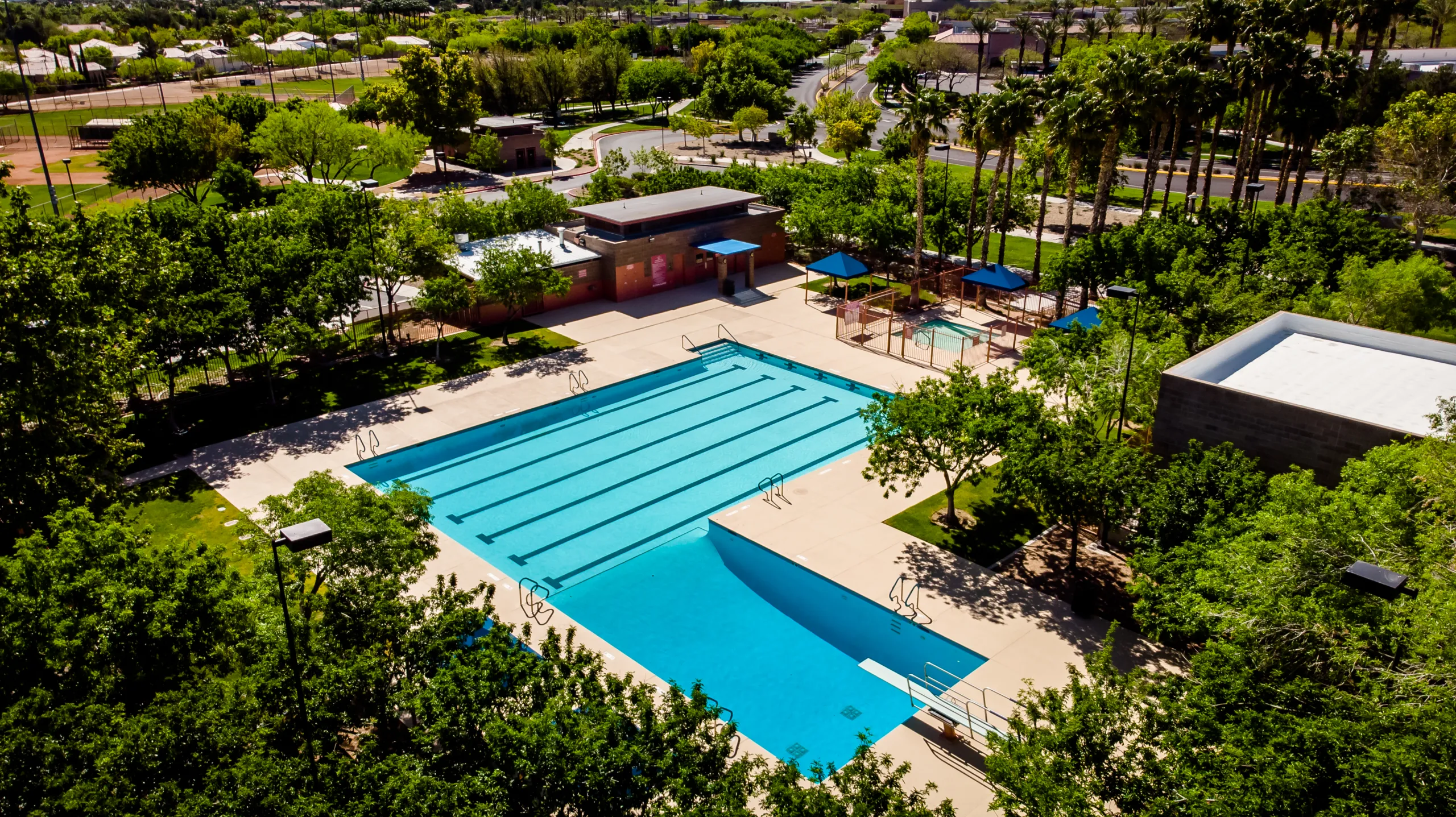 Aerial view of an outdoor swimming pool surrounded by trees, with lounge chairs, shaded seating areas, and a nearby building. The pool has swim lanes and bright blue water. The setting is sunny and well-maintained.