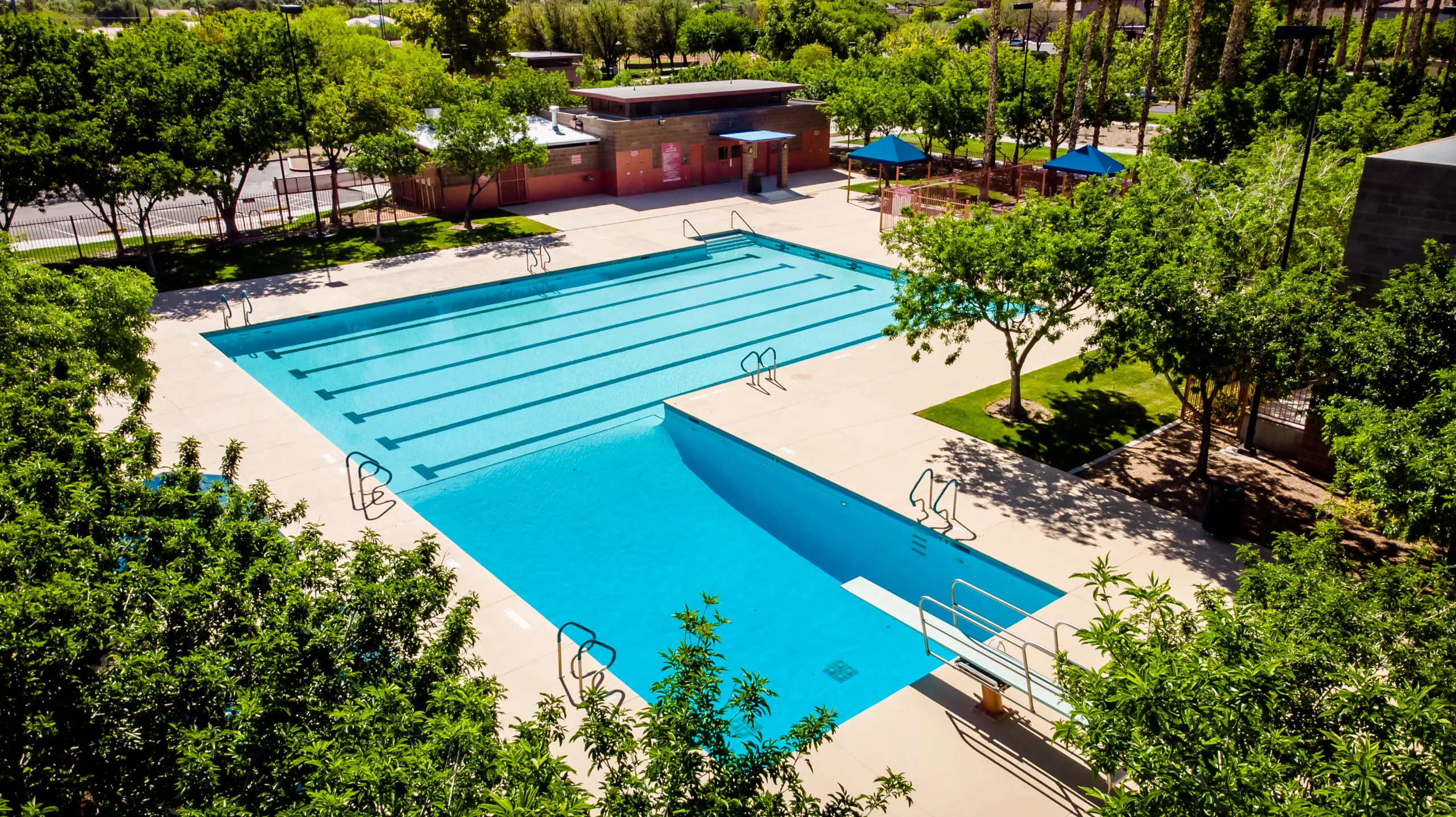A large outdoor swimming pool, surrounded by trees and shaded areas, with clear blue water, pool ladders, and a building with changing rooms in the background. The pool deck is empty and the sky is sunny.