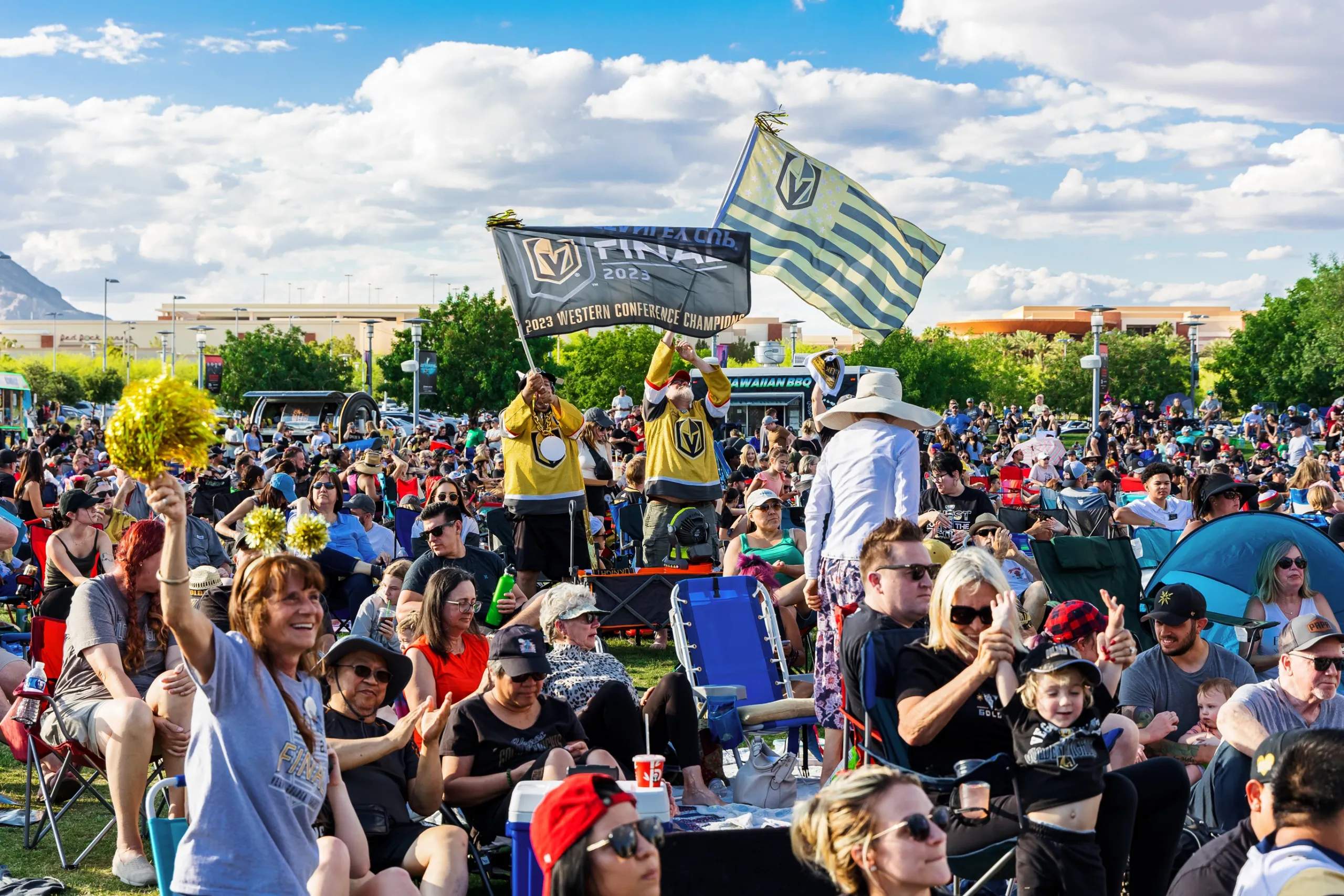 2023 VGK Watch Party A large crowd gathers outdoors at a lively event, with people sitting on lawn chairs and smiling. In the background, fans wave Vegas Golden Knights flags. The sky is partly cloudy, and mountains are visible in the distance.