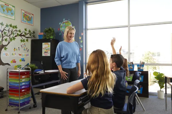 A teacher smiles at two students seated at desks, one raising their hand, in a bright classroom decorated with maps, colorful bins, and a tree mural.