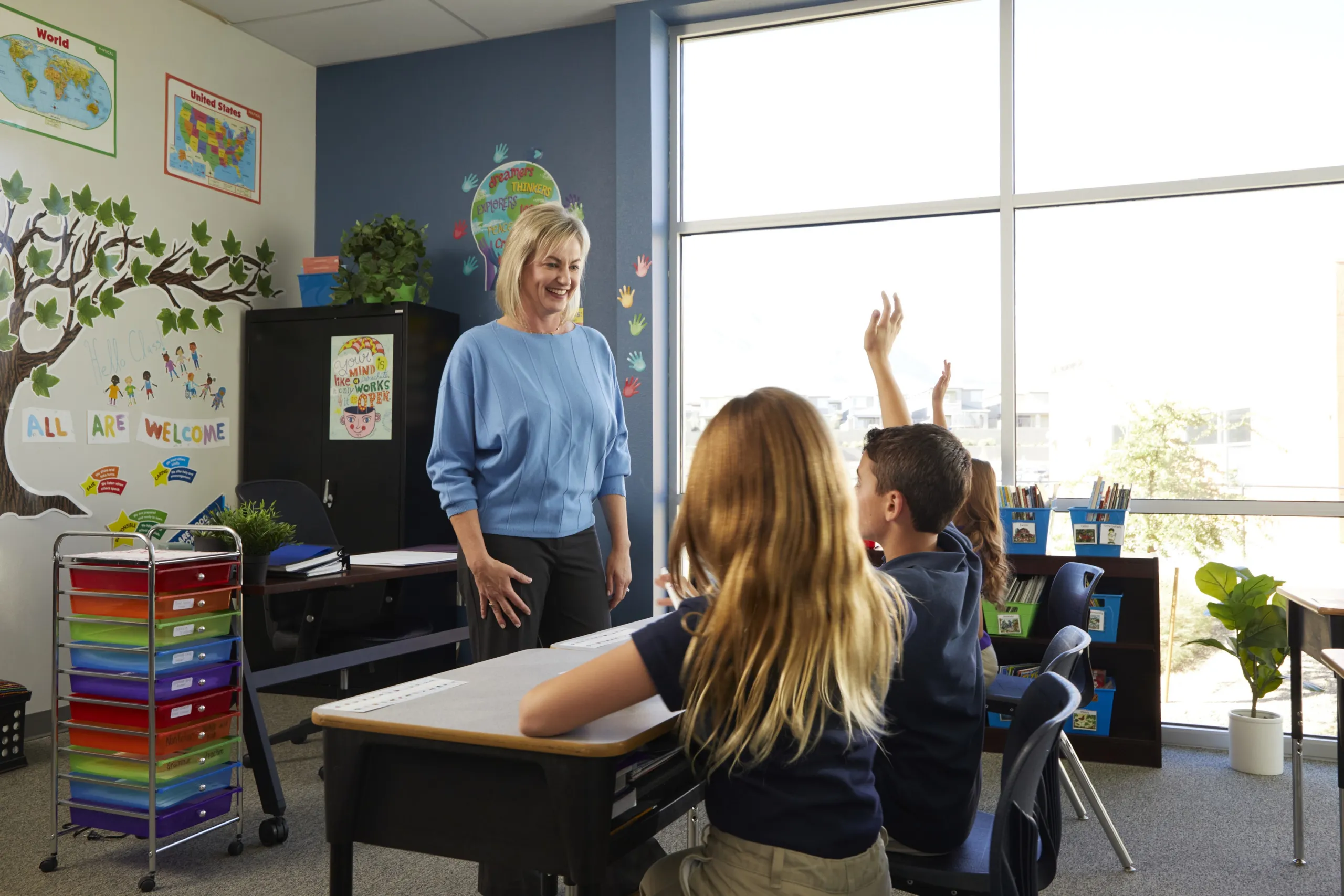 A teacher smiles at two students seated at desks, one raising their hand, in a bright classroom decorated with maps, colorful bins, and a tree mural.