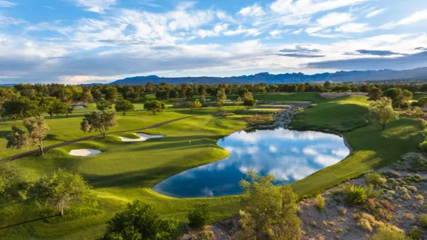 Aerial view of a lush golf course with sand traps, a reflective pond, trees, and distant mountains under a blue sky with scattered clouds.