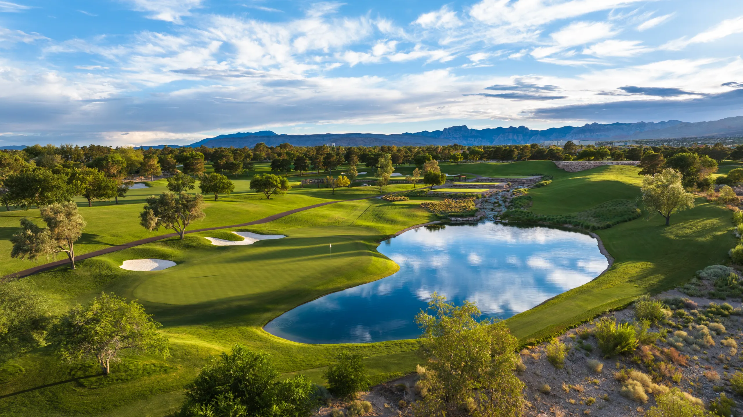 Aerial view of a lush golf course with sand traps, a reflective pond, trees, and distant mountains under a blue sky with scattered clouds.
