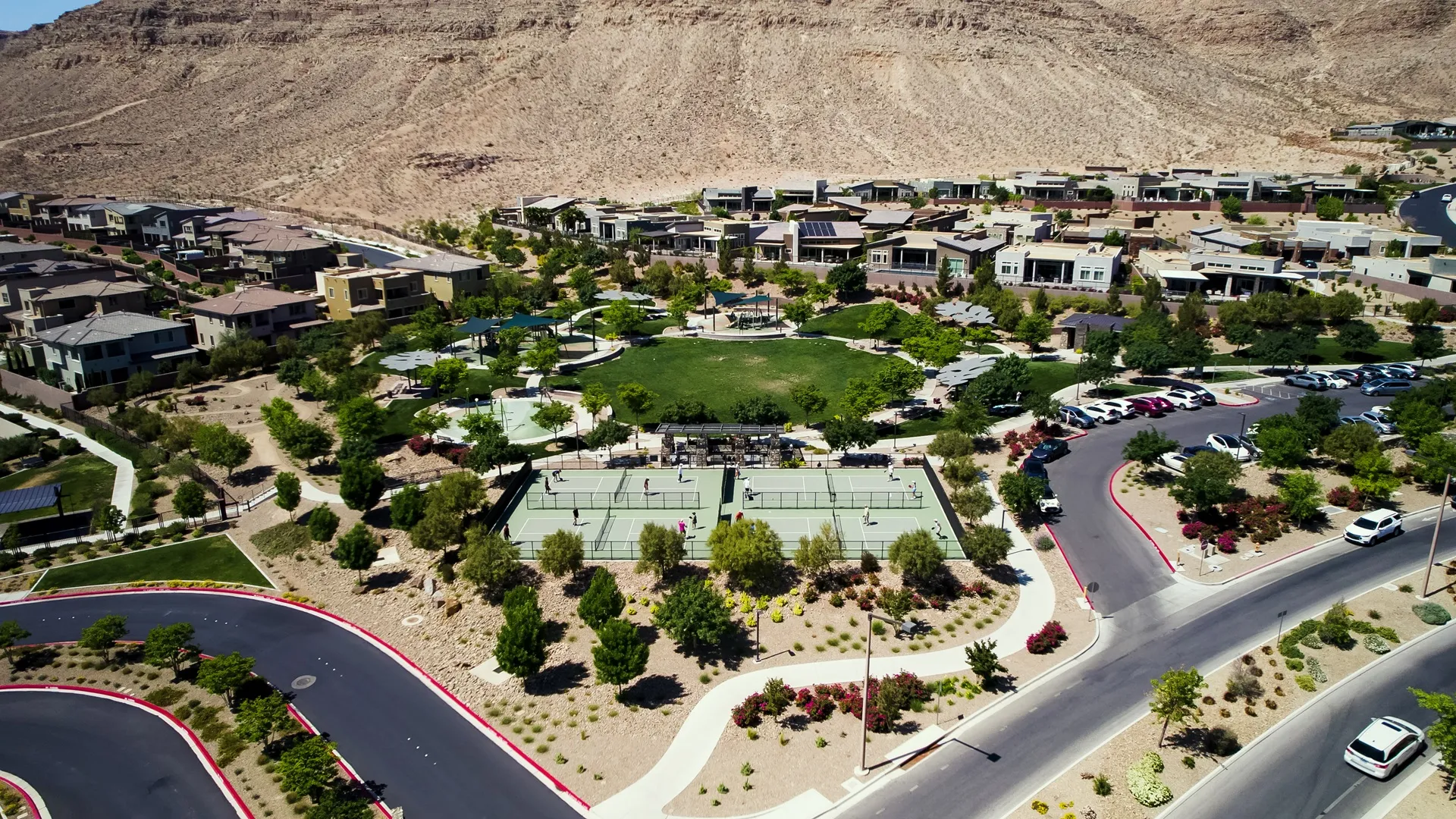 Aerial view of a suburban neighborhood with houses, tennis courts, a green park, trees, and parked cars, set against a backdrop of rocky hills.