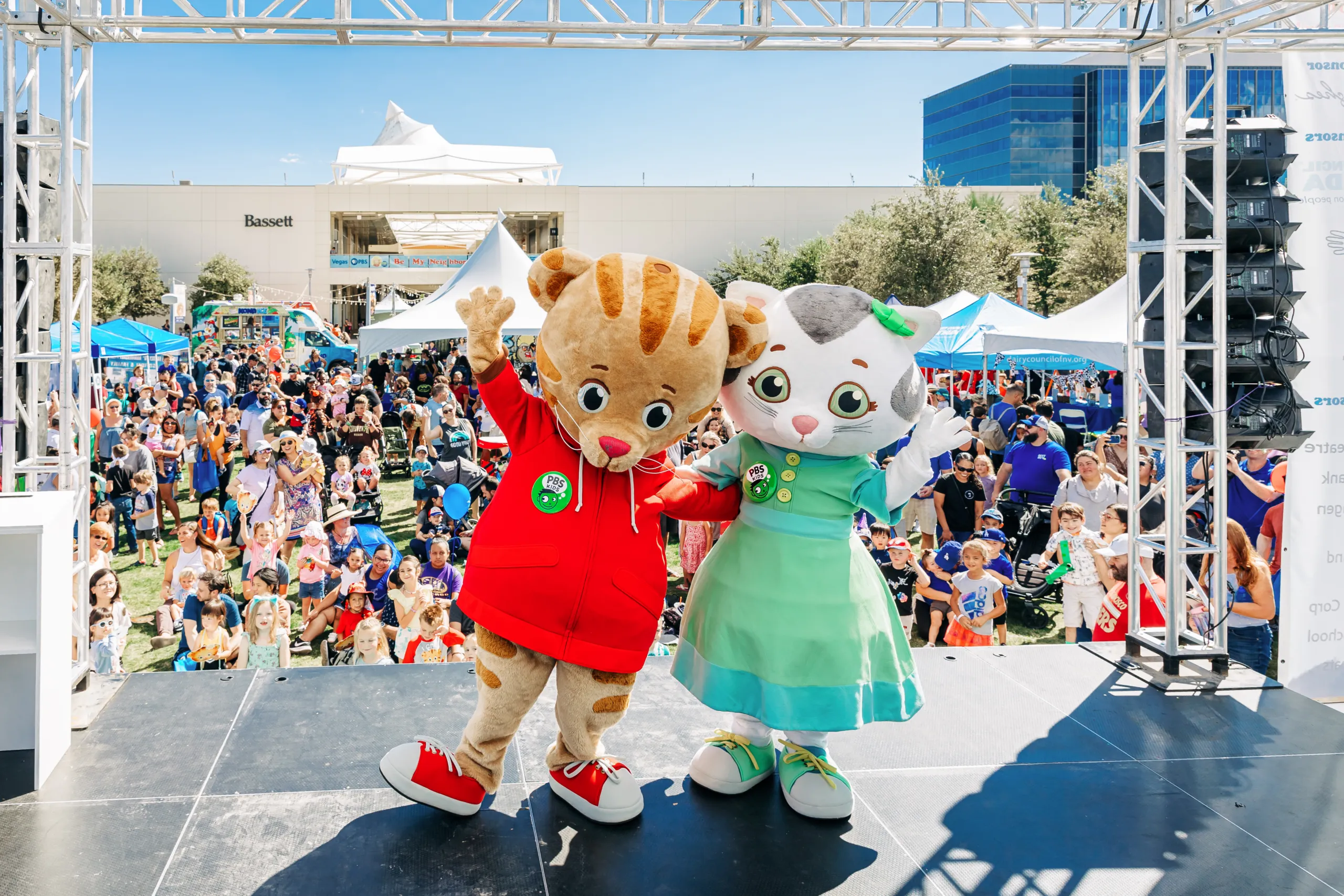 Two costumed characters, a tiger in a red hoodie and a cat in a green dress, stand on a stage waving to a large outdoor crowd at a family event with tents and booths in the background.