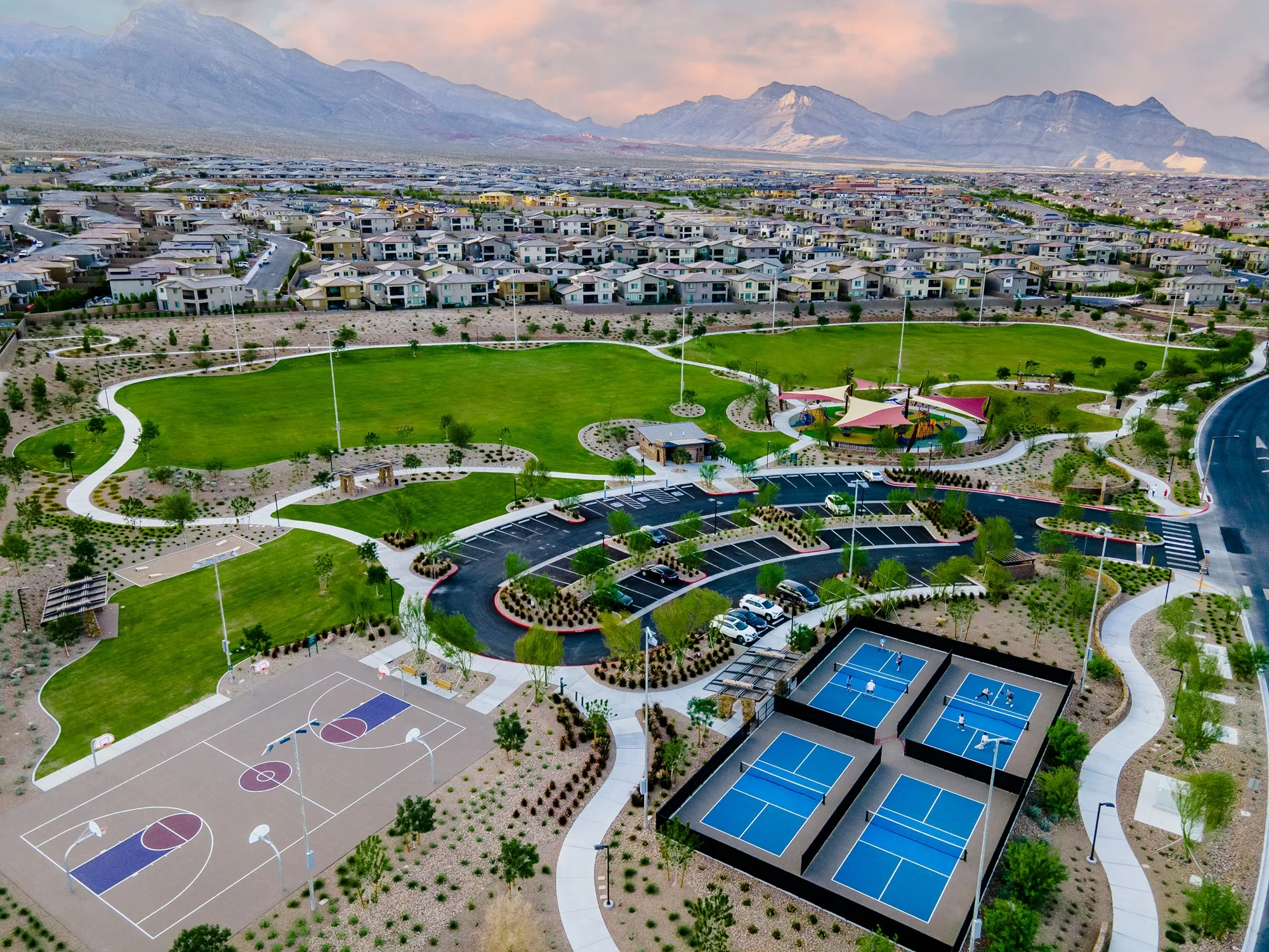 Aerial view of a suburban park with green lawns, basketball courts, tennis or pickleball courts, playgrounds, walking paths, parking lot, and surrounding houses, with mountains in the background.
