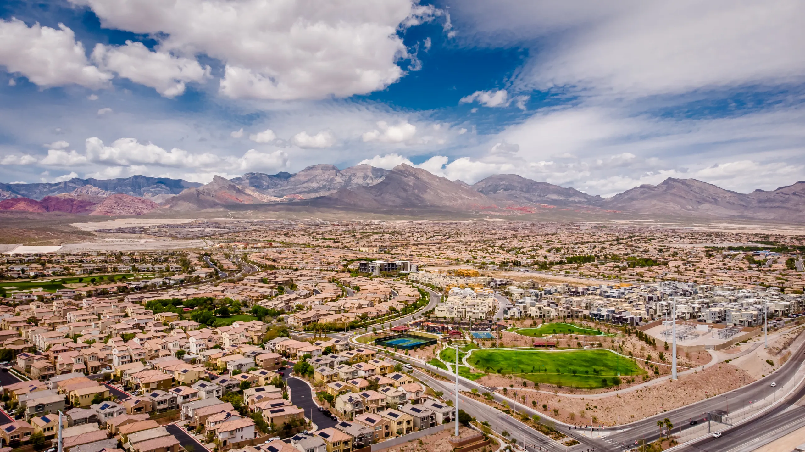 Aerial view of a sprawling suburban neighborhood with many houses, green parks, and roads, set against a backdrop of mountains under a partly cloudy sky.