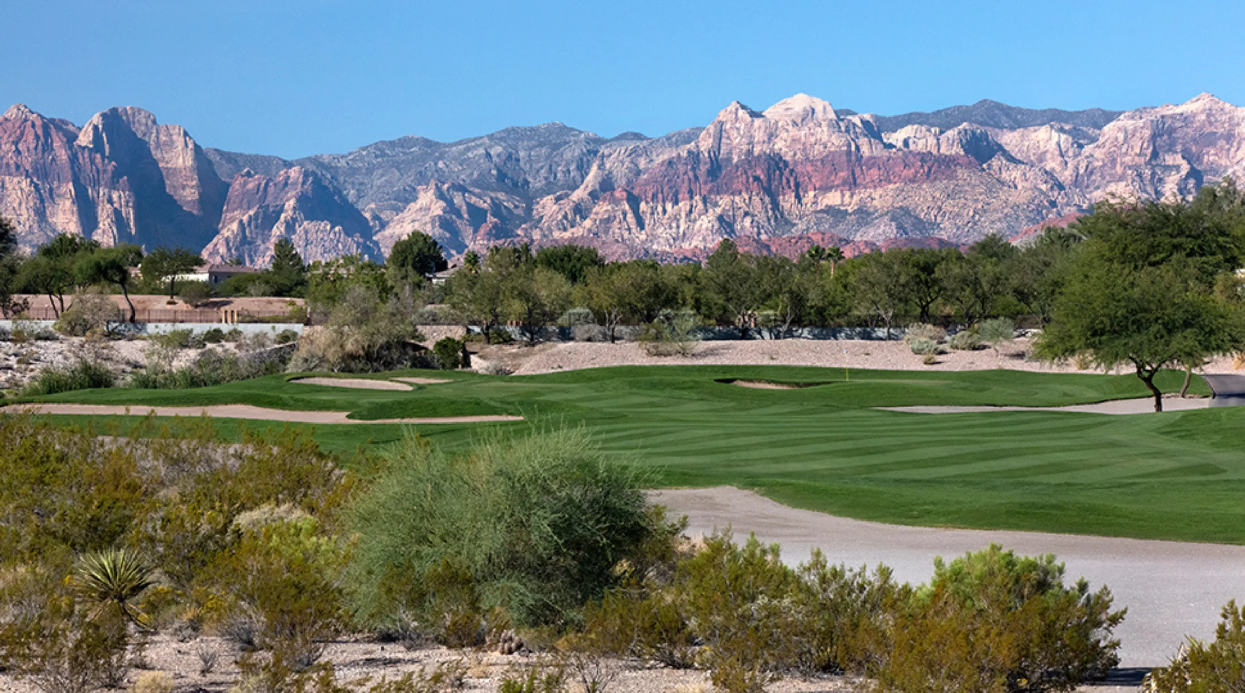 A lush green golf course with sand traps in the foreground, desert plants surrounding it, and colorful rocky mountains under a clear blue sky in the background.